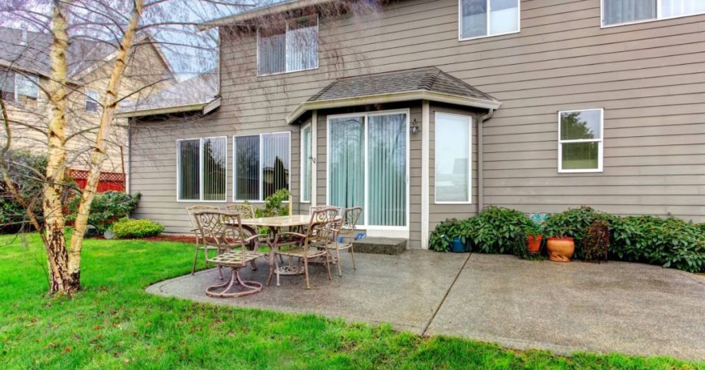 a backyard view of a modern suburban house with beige siding and large windows. The patio features a metal table and chairs set, surrounded by green grass, shrubs, and a bare birch tree. 