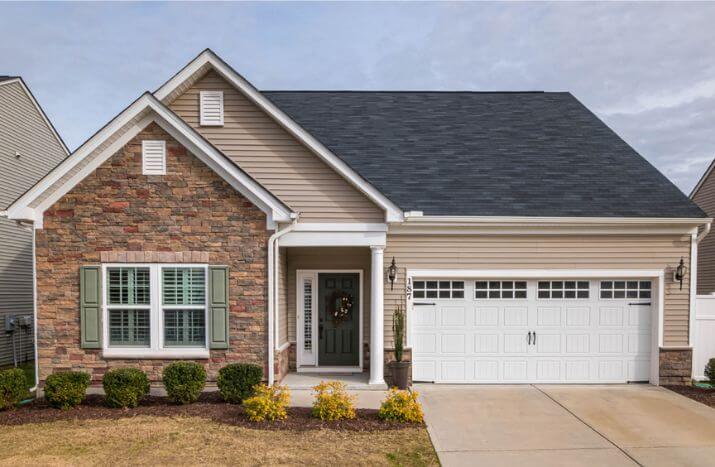 A modern suburban home with a light-colored stone and beige siding exterior, featuring a black shingle roof, a white garage door, and a green front door with side shutters. The driveway and small landscaped front yard are visible.